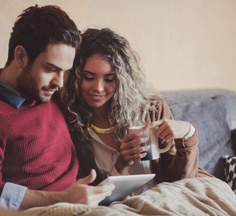 A male sits on a couch with his arm around his smiling girlfriend who holds a cup of coffee in her hands and both are underneath a blanket watching Better Together marriage preparation videos