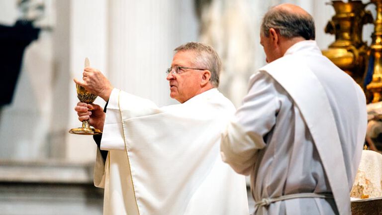 A catholic priest in all white cloaks holds up a chalice and eucharist in his hands during Sunday mass while a deacon stands next to him with his eyes closed and head down