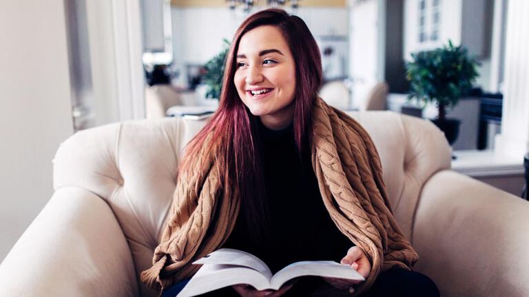 A smiling teenage girl sits on a tan couch in her living room while reading through Dynamic Catholic's confirmation program, Decision Point