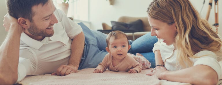 A smiling father and mother both wearing white shirts sit on the carpet in their home between their newborn girl who lays on her stomach with her head held up 