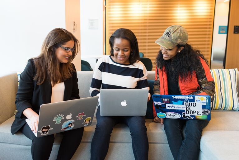 Three college aged women sit on a couch in the library together each holding their laptops watching a Sunday Gospel reflection video. 