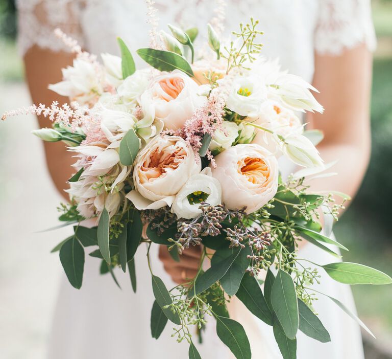 A large bridal bouquet with green leafs, white roses and blush flowers are held in the front of the camera by a bride in her wedding dress 