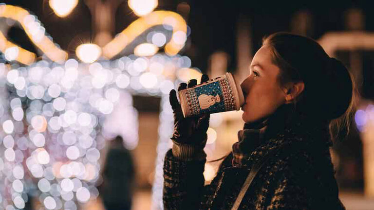 Woman drinks a cup of coffee. Christmas lights are hung in street behind her. 