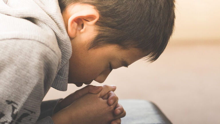 A young boy leans over a chair praying with his hands folded in front and his eyes closed as he prepares for first communion